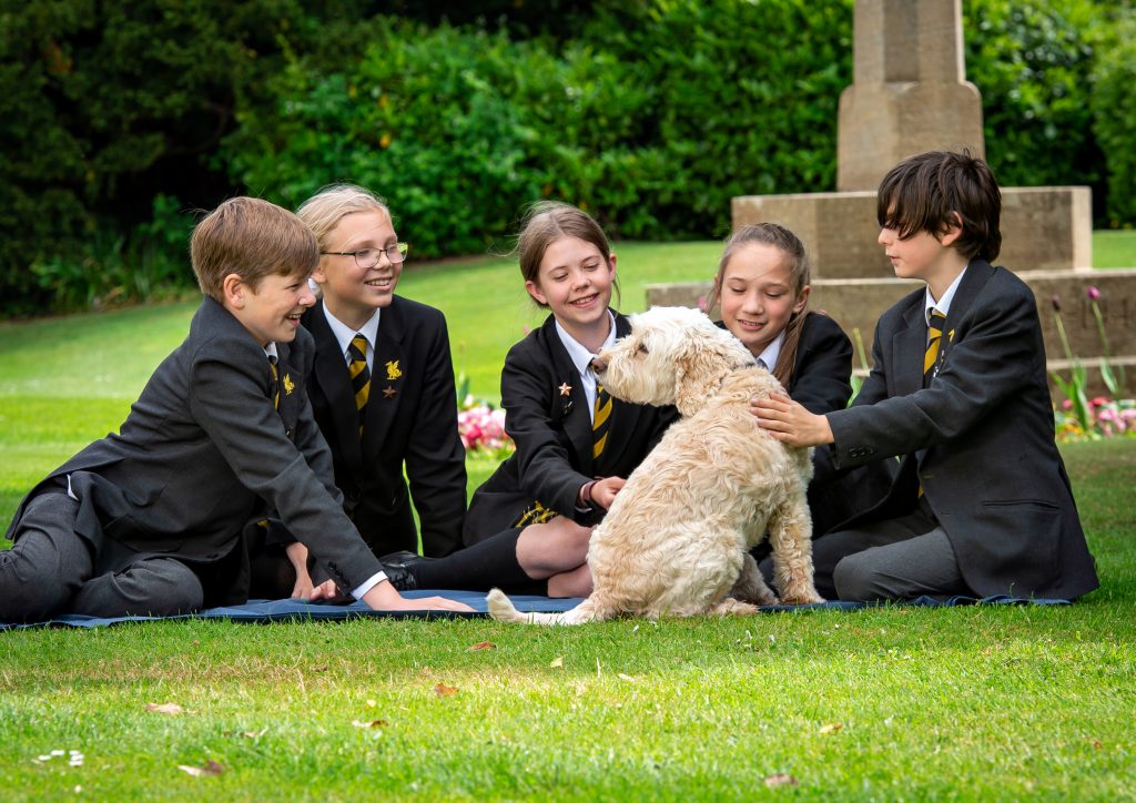 5 students wearing school uniform sat on the grass petting a dog